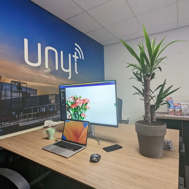  Bureausamenstelling met een Macbook, monitor, iPhone, muis, beker en een plant, wat het dynamische en efficiënte werkomgeving bij UNYT symboliseert.